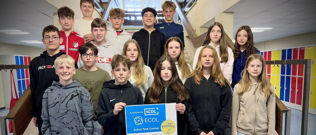 Eine Gruppe von Schülern, die auf einer Treppe stehen und ein blaues Zertifikat für die ICDL-Prüfung halten. Einige tragen Jacken, Brillen und Turnschuhe. Sie posieren für ein Foto.