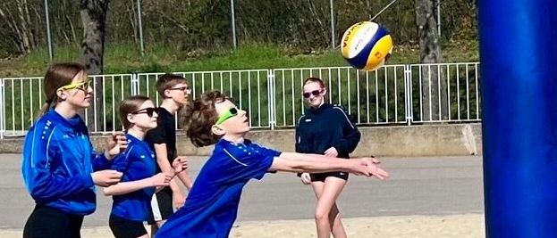 Eine Gruppe junger Leute spielt Volleyball auf einem sandigen Platz. Ein Spieler ist in der Luft und bereitet sich darauf vor, den Ball zu schlagen. In der Nähe sitzt eine Person auf einer Bank und beobachtet das Spiel.