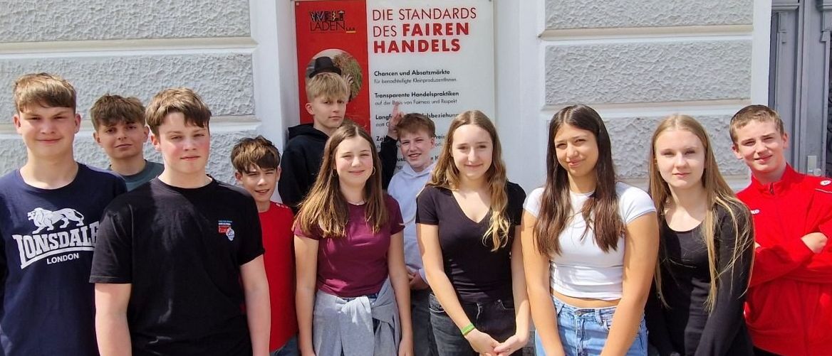 Eine Gruppe junger Menschen posiert vor einem Gebäude mit einem roten Schild, das Die Standards des Fairen Handels besagt.