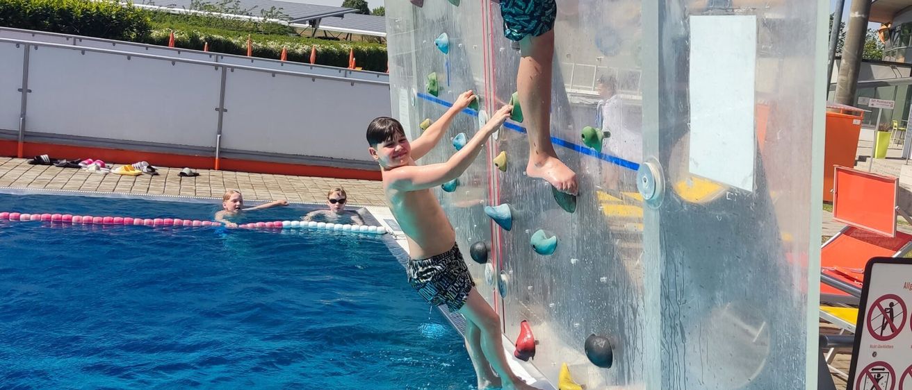 Ein Junge klettert an einer aufblasbaren Kletterwand in einem Schwimmbad, während im Hintergrund zwei Personen schwimmen.