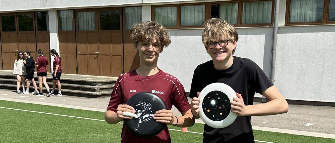 Zwei junge Männer halten Frisbees vor einem Schulgebäude. Einer hat ein schwarzes Frisbee mit Sonnensystem-Design und der andere hält ein weißes.