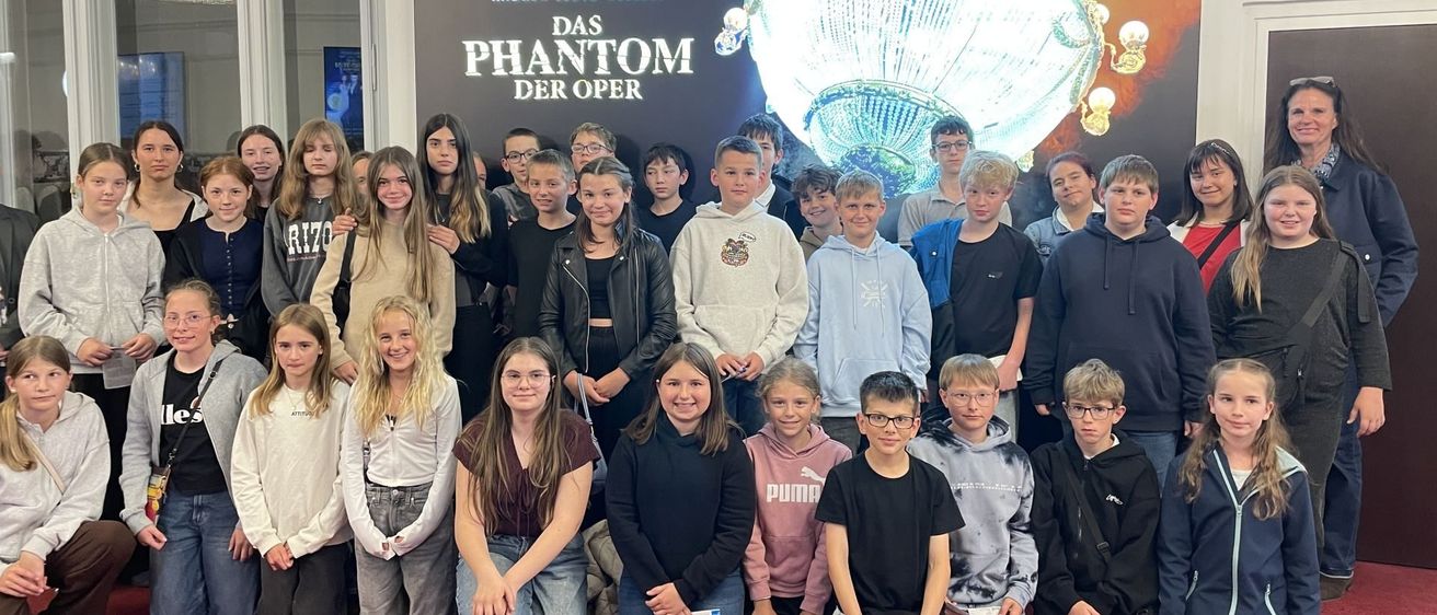 Eine Gruppe von Kindern posiert vor einem Werbeplakat für das Musical 'Das Phantom der Oper'. Sie lächeln und scheinen Spaß zu haben.