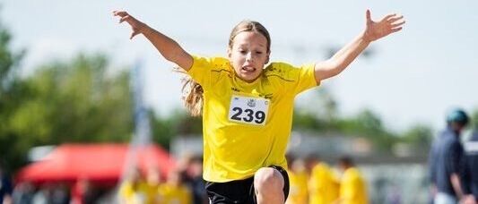 Ein junges Mädchen in einem gelben Shirt springt während eines Leichtathletik-Events in den Sand. Andere Athleten beobachten hinter ihr.