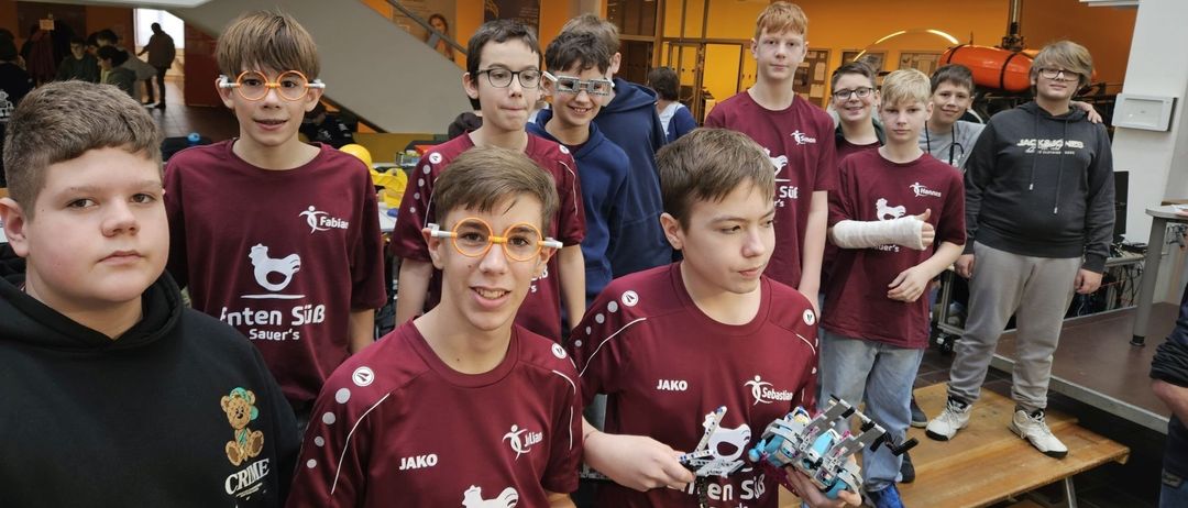 Mehrere Jungen in roten Trikots und mit Brille stehen zusammen und lächeln. Sie scheinen Schüler zu sein, die an einem Roboterwettbewerb teilnehmen. Ein Junge hält einen Roboter in beiden Händen.