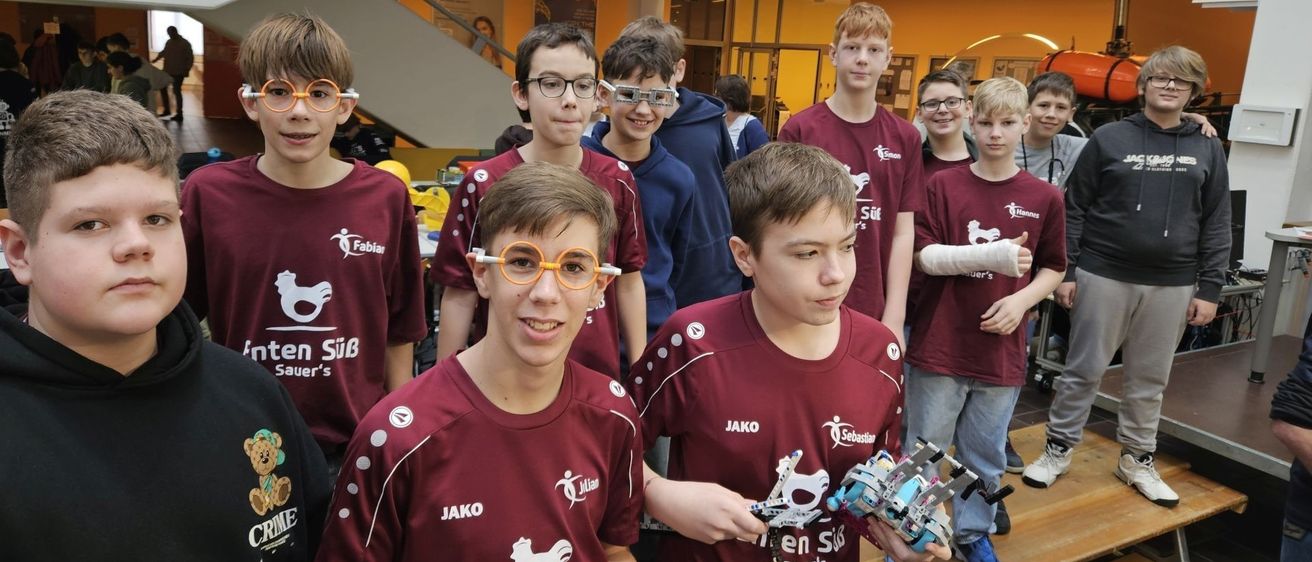 Mehrere Jungen in roten Trikots und mit Brille stehen zusammen und lächeln. Sie scheinen Schüler zu sein, die an einem Roboterwettbewerb teilnehmen. Ein Junge hält einen Roboter in beiden Händen.