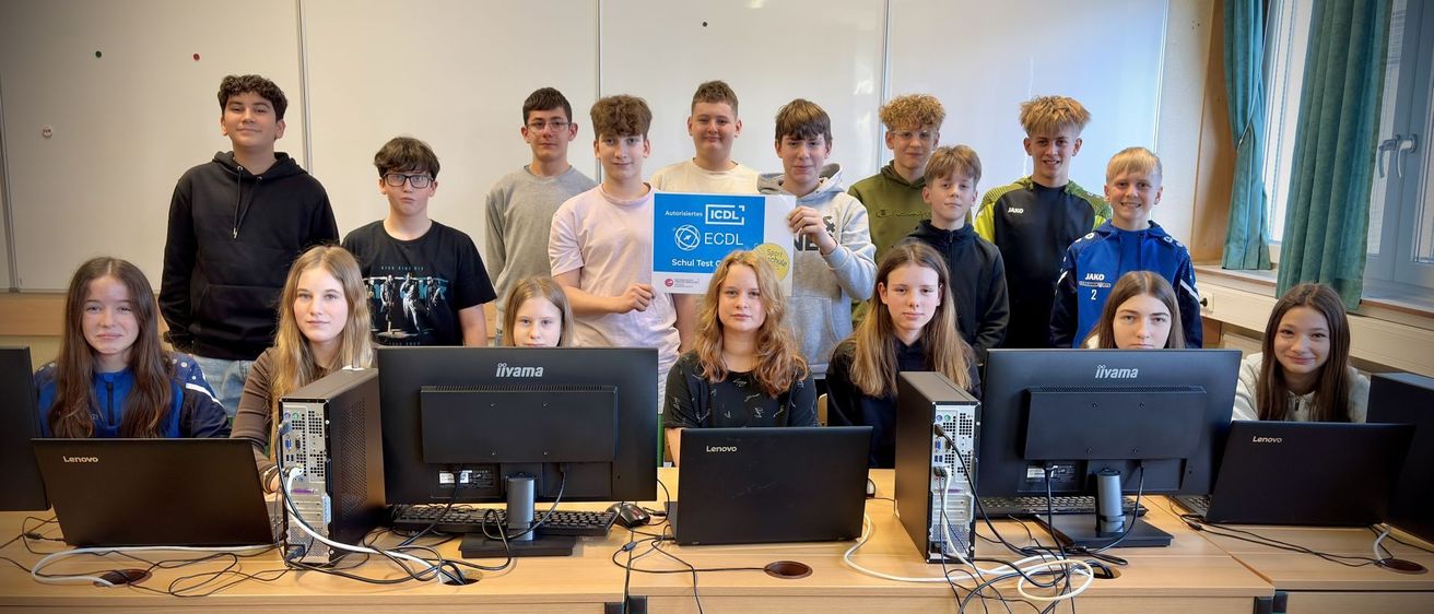 Eine Gruppe junger Schüler in einem Klassenzimmer, die für ein Foto mit mehreren Computern posieren. Ein blauer Banner in der Mitte trägt den Text 'ECD L Schul Test'.