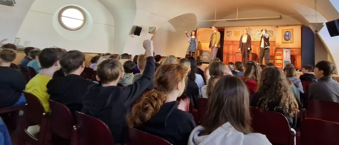 Mehrere Personen sitzen in einem Auditorium. Einige beobachten eine Aufführung auf der Bühne mit stehenden Personen, die auftreten. Scheinwerfer sind an der Decke.
