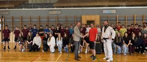 Eine Gruppe von Menschen posiert für ein Foto in einer Turnhalle. Zwei Männer schütteln sich die Hände in der Mitte. Sie sind von Schülern umgeben. Die Turnhalle hat Holzböden und Tribünen.