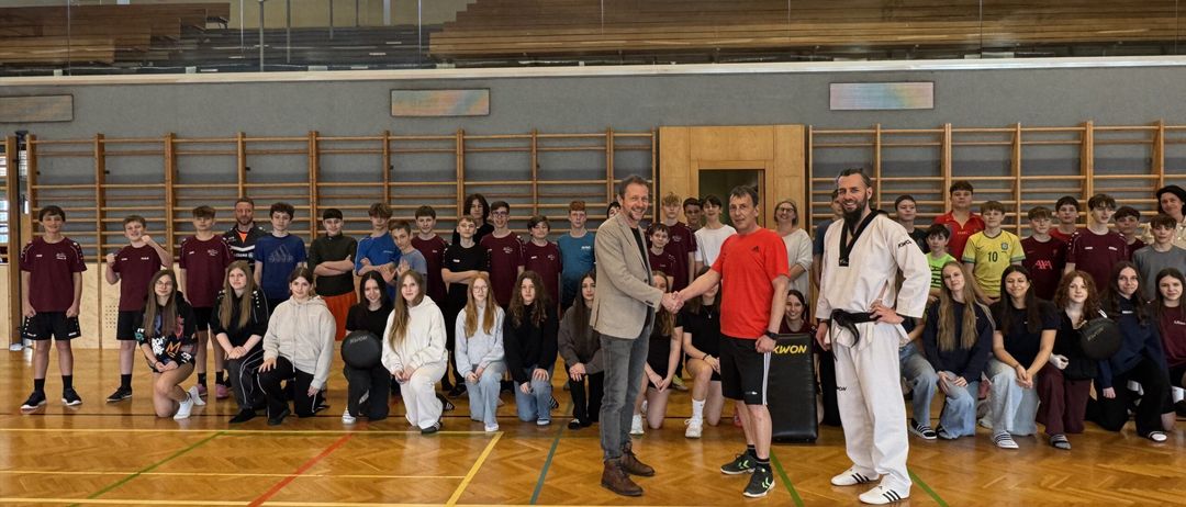 Eine Gruppe von Menschen posiert für ein Foto in einer Turnhalle. Zwei Männer schütteln sich die Hände in der Mitte. Sie sind von Schülern umgeben. Die Turnhalle hat Holzböden und Tribünen.