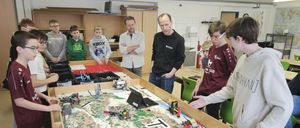 In einem Klassenzimmer versammeln sich mehrere Schüler um einen großen Tisch mit einer Lego-gebauten Landschaft. Zwei Männer in Jacken beobachten die Schüler. Der Tisch hat verschiedene Lego-Strukturen, darunter einen Kran und eine Brücke.