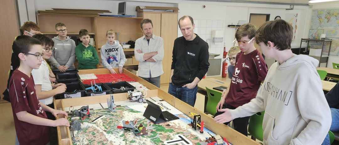 In einem Klassenzimmer versammeln sich mehrere Schüler um einen großen Tisch mit einer Lego-gebauten Landschaft. Zwei Männer in Jacken beobachten die Schüler. Der Tisch hat verschiedene Lego-Strukturen, darunter einen Kran und eine Brücke.