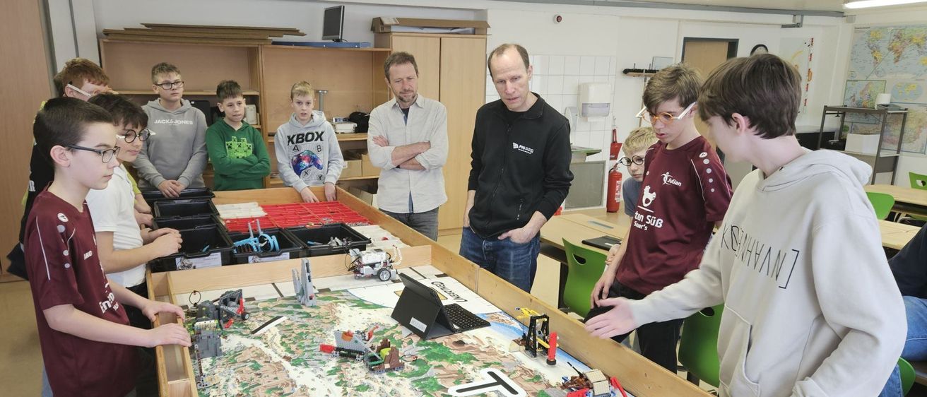 In einem Klassenzimmer versammeln sich mehrere Schüler um einen großen Tisch mit einer Lego-gebauten Landschaft. Zwei Männer in Jacken beobachten die Schüler. Der Tisch hat verschiedene Lego-Strukturen, darunter einen Kran und eine Brücke.