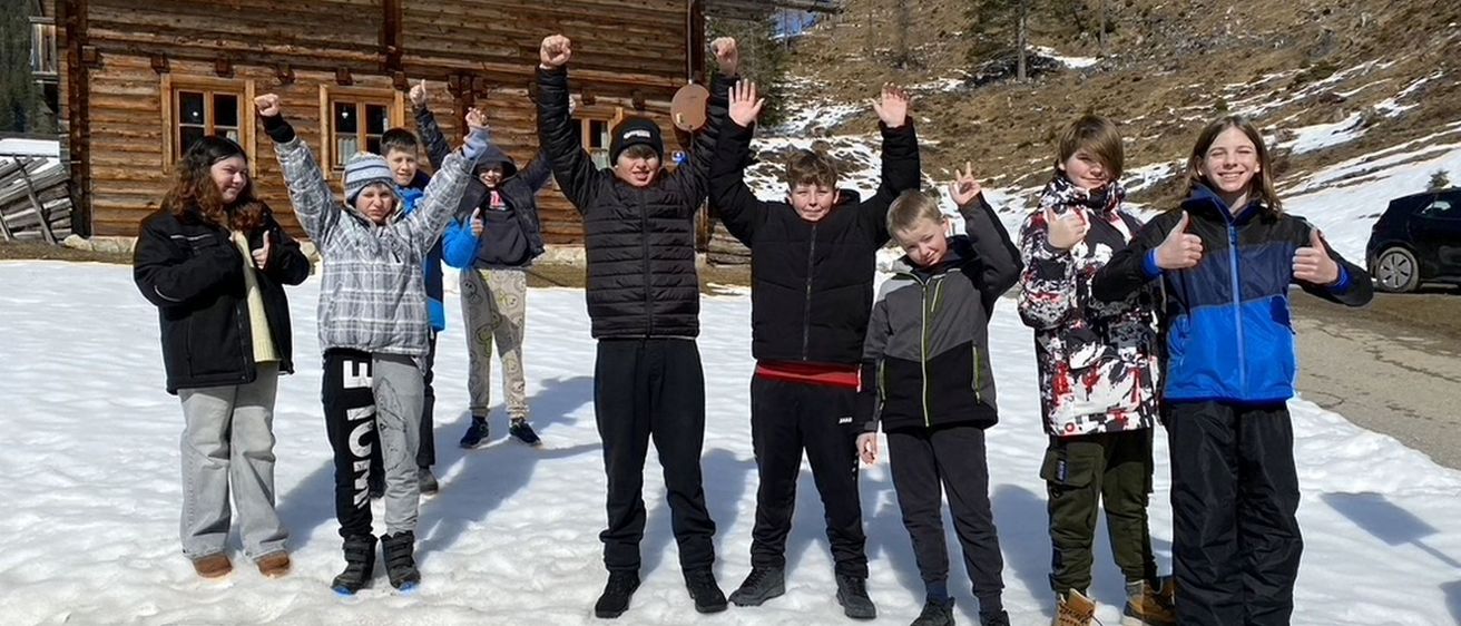 Eine Gruppe von Kindern posiert für ein Foto im Schnee vor einem Holzhaus. Sie tragen Winterkleidung.