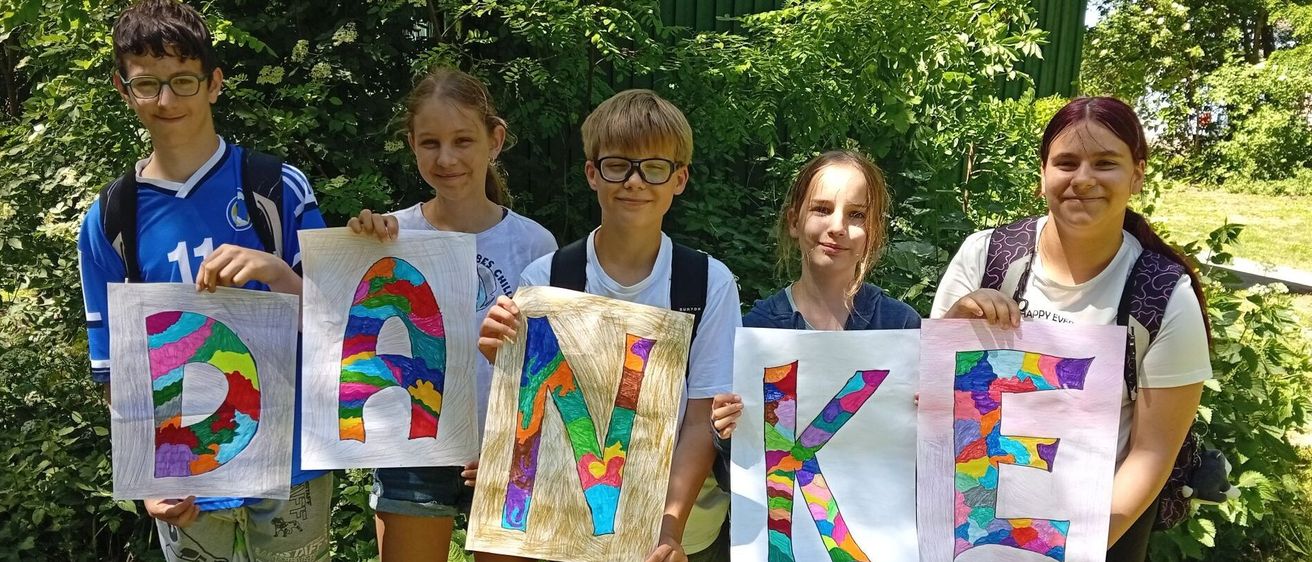 Fünf Kinder stehen in einer Reihe und halten große bunte Buchstaben hoch, die das Wort DANKE bilden. Jedes Kind hält einen anderen Buchstaben und lächelt für die Kamera. Sie sind von Grün umgeben.