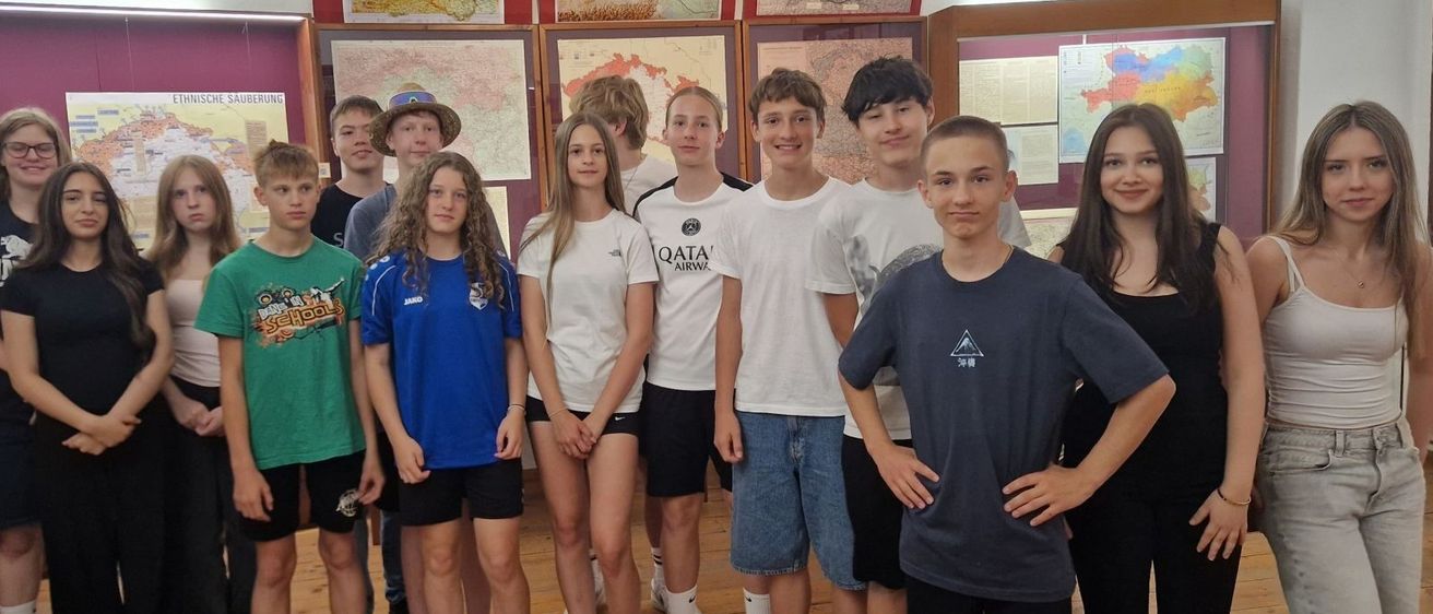 Eine Gruppe junger Leute in Freizeitkleidung steht vor einem Raum mit Karten und Dokumenten zur Ausstellung. Sie lächeln und posieren für ein Foto.
