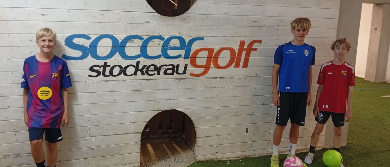Drei Kinder stehen auf einem grünen Kunstrasen und posieren vor einer Wand mit dem Wort soccergolf. Zwei Jungen tragen blaue und rote Trikots.
