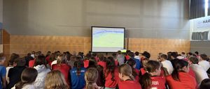 Ein Klassenzimmer voller Schüler, die rote Trikots tragen, beobachten ein Fußballspiel auf einer Projektionsleinwand.