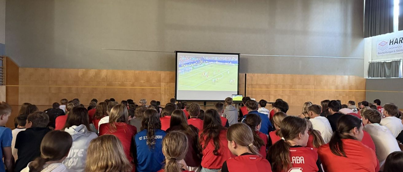 Ein Klassenzimmer voller Schüler, die rote Trikots tragen, beobachten ein Fußballspiel auf einer Projektionsleinwand.