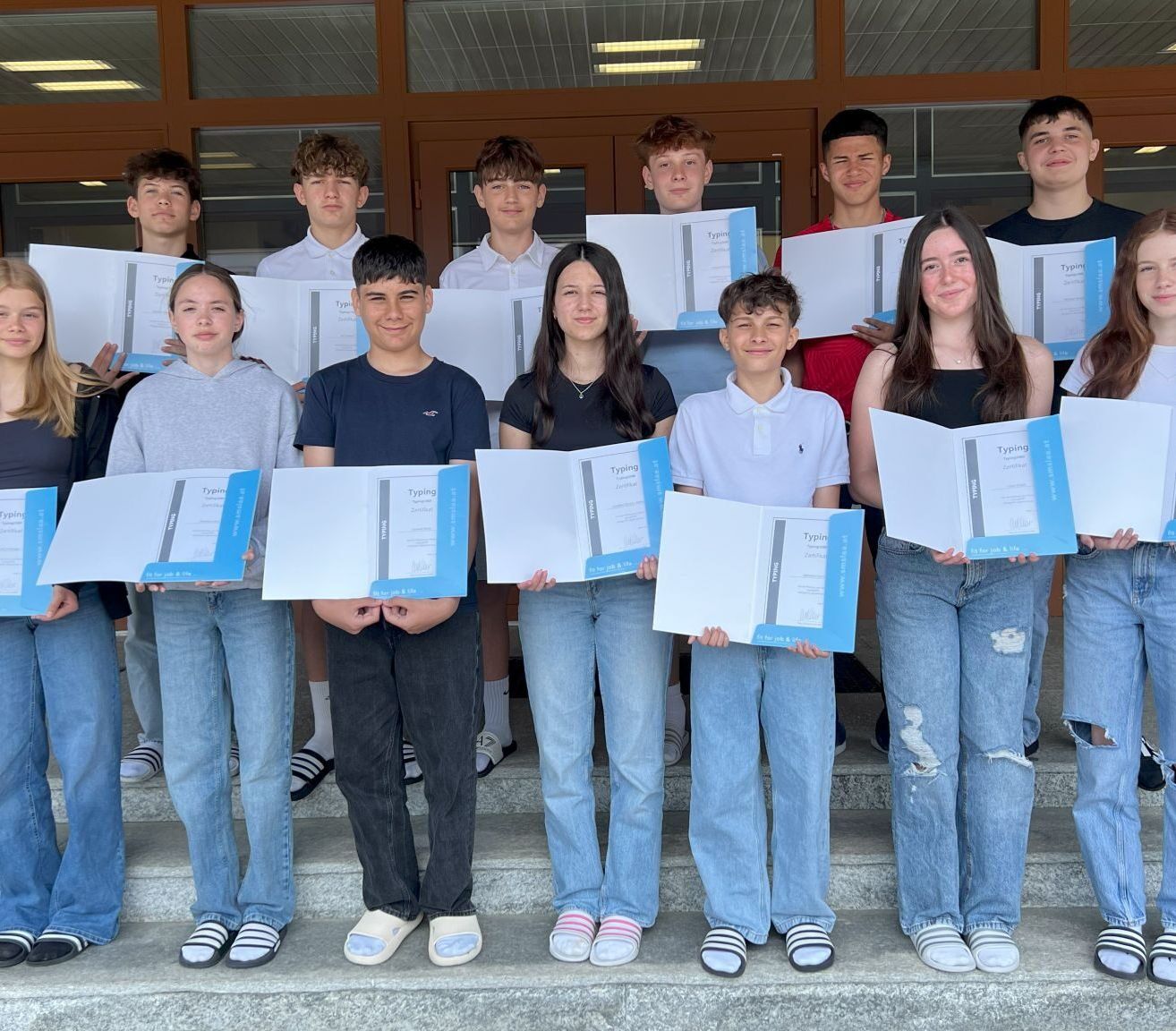 Eine Gruppe junger Schüler steht auf Stufen, lächelt und hält Zertifikate. Sie tragen Freizeitkleidung und Turnschuhe. Der Raum hat Holztüren und helle Beleuchtung.