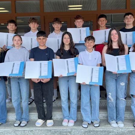 Eine Gruppe junger Schüler steht auf Stufen, lächelt und hält Zertifikate. Sie tragen Freizeitkleidung und Turnschuhe. Der Raum hat Holztüren und helle Beleuchtung.