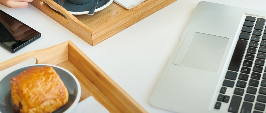 Eine Person sitzt an einem Tisch mit Laptop und hält eine Kaffeetasse. Tabletts mit Croissants und Gebäck, ein Telefon und ein Buch sind in der Nähe. Der Text lautet 'Kostenloser Online-Frühstücks-Talk'.