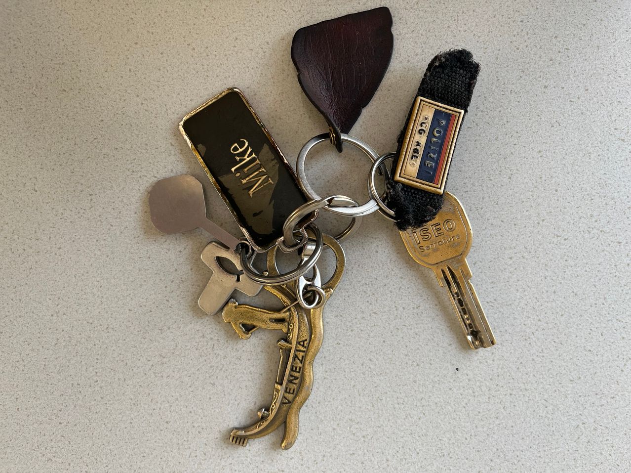 Eine Sammlung von Schlüsseln an einem Metallring, einschließlich einer Löwenfigur, eines Kreuzes, eines Löffels und eines venezianischen Tags. Ein Schlüssel mit schwarzem Griff und den Worten 'Polizei' ist ebenfalls enthalten.