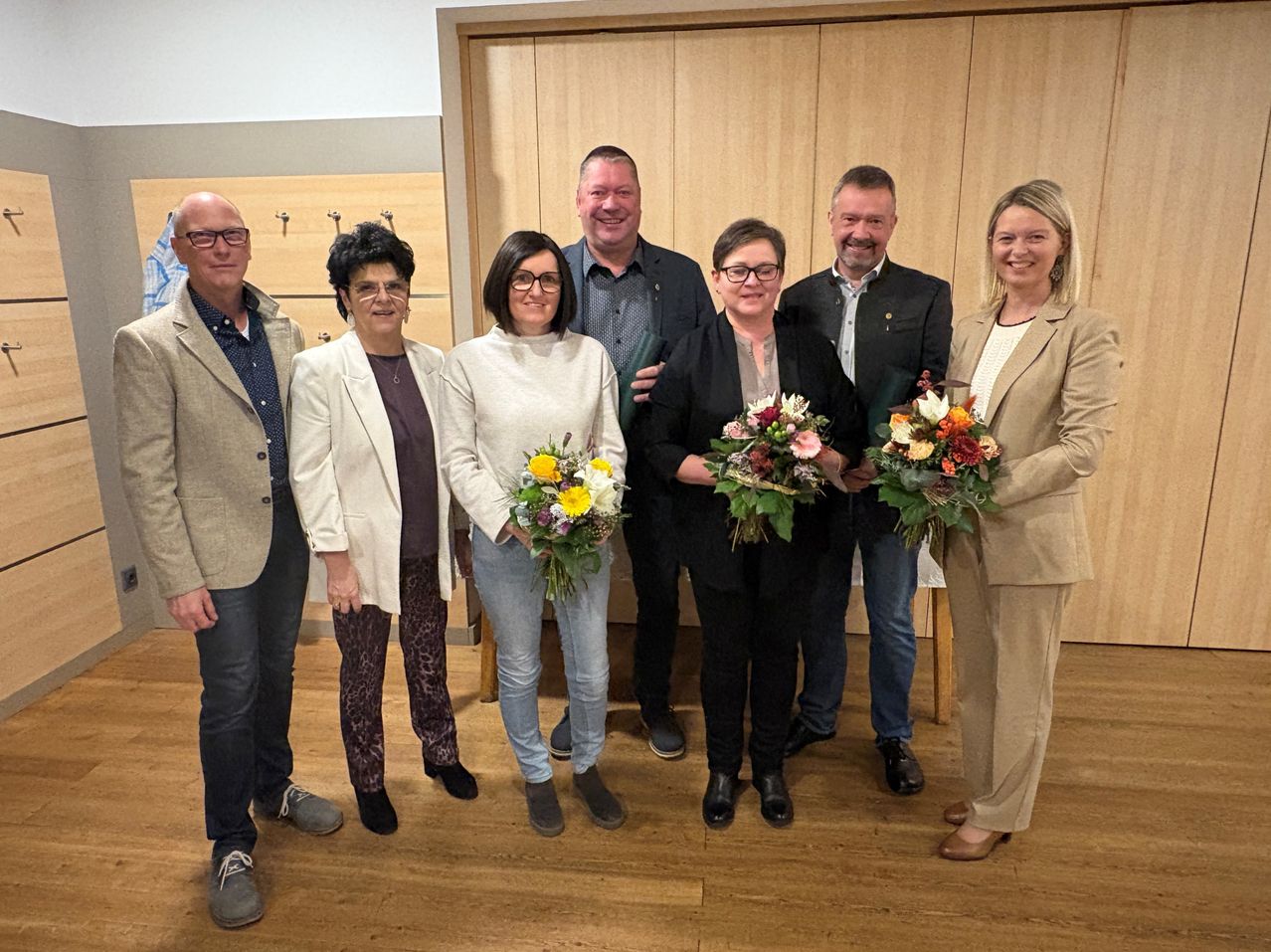 Sechs Personen stehen zusammen und halten Blumen. Hinter ihnen dient eine Holzwand als Hintergrund. Sie alle scheinen für die Kamera zu lächeln.