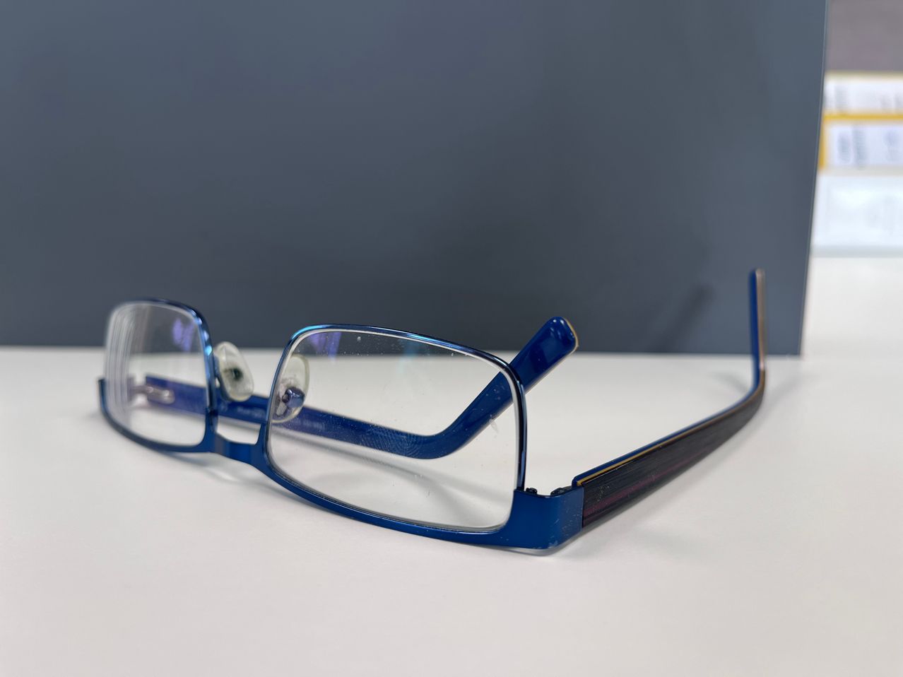 Blue-framed glasses with clear lenses rest on a white surface, reflecting a gray background.