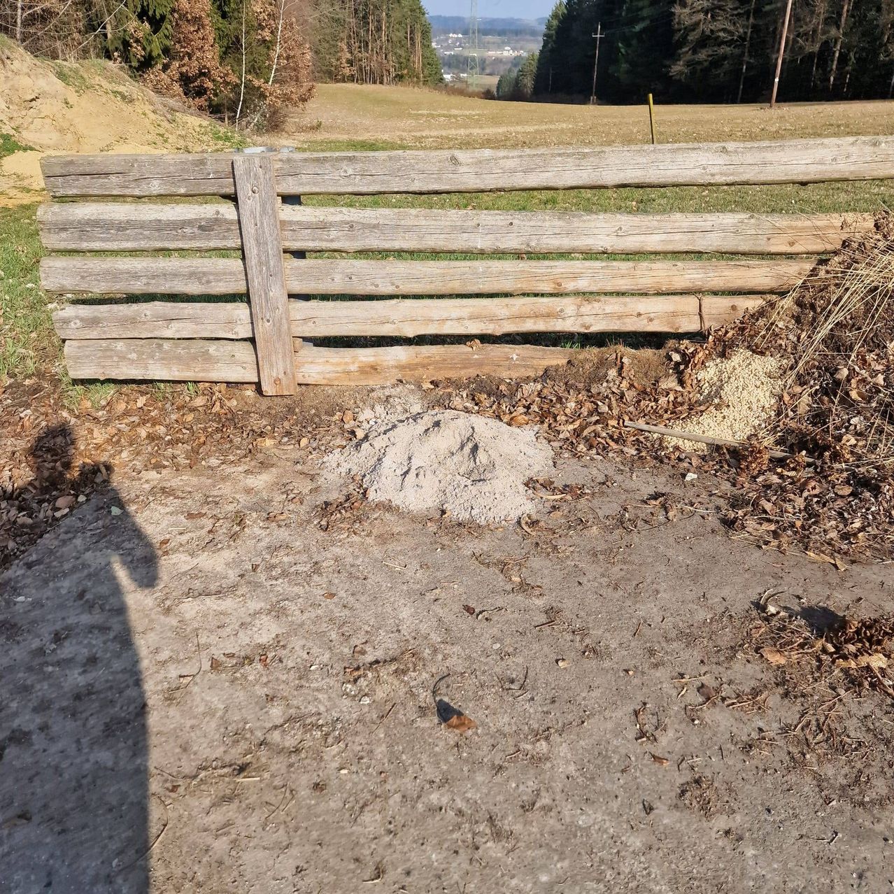 Ein Holzzaun steht neben einem Erdweg mit einem Haufen Laub und Kies. Im Hintergrund zeigt sich ein Rasenfeld mit Bäumen und einer Fernsicht auf Gebäude.