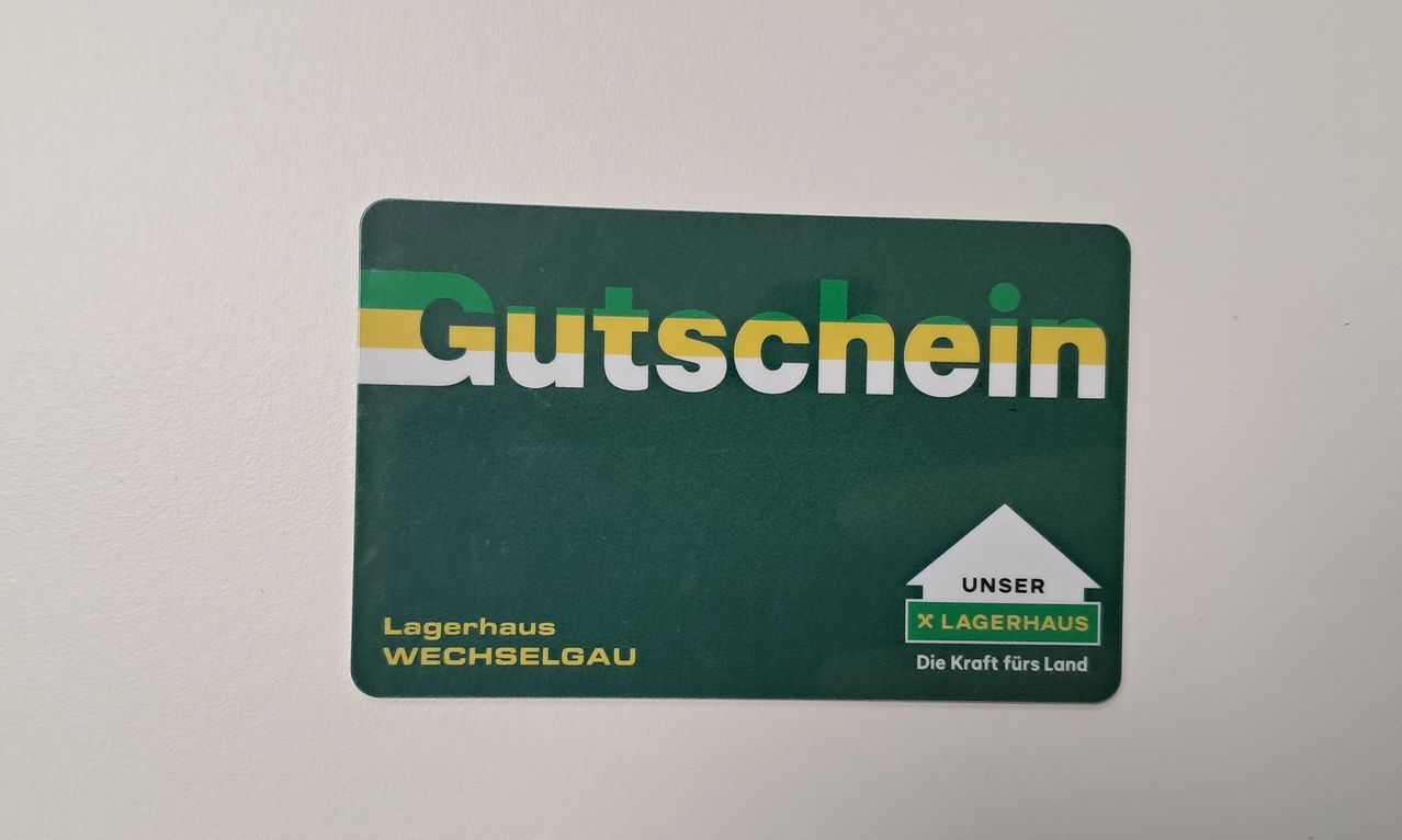 Ein grüner Gutschein für Lagerhaus Wechselgau ist auf einem weißen Hintergrund zu sehen. Er zeigt ein weißes Haussymbol mit dem Text 'Unser Lagerhaus'. Darunter steht 'Die Kraft fürs Land'.