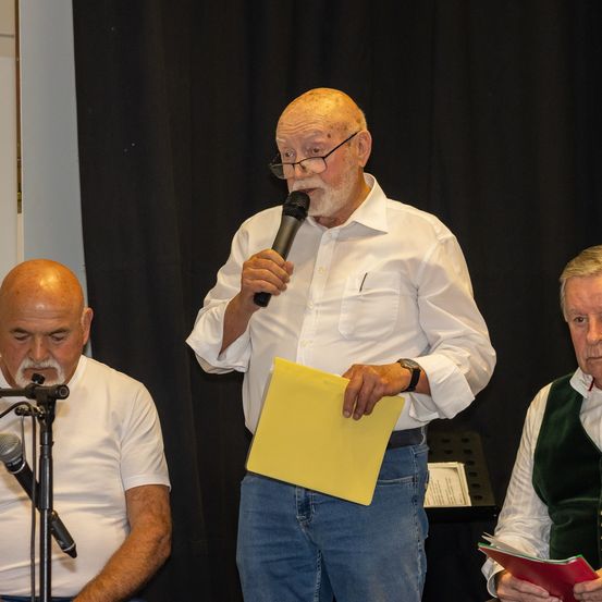 Ein älterer Mann spricht in ein Mikrofon vor einem schwarzen Vorhang. Zwei andere Männer sitzen neben ihm, einer mit einem Buch und der andere mit einem Notizblock.