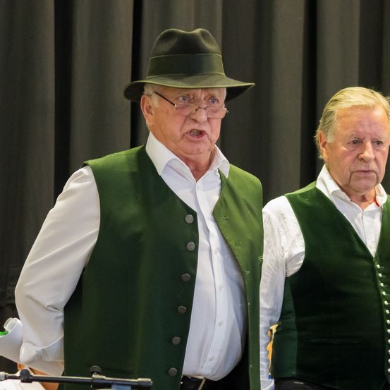 Zwei ältere Männer stehen nebeneinander und tragen grüne Westen, weiße Hemden und Gürtel. Der Mann auf der linken Seite trägt einen Hut und eine Brille. Sie scheinen zu singen oder aufzutreten.