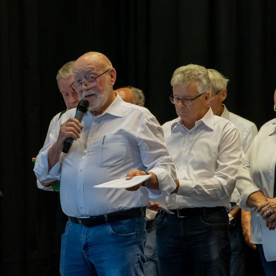 Ein älterer Mann mit Brille hält ein Mikrofon und Papier in der Hand und steht vor einer Gruppe älterer Personen, darunter eine mit einer Uhr, vor einem schwarzen Hintergrund.