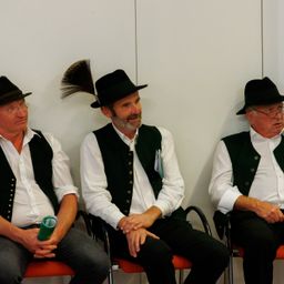 Drei ältere Männer in traditioneller Kleidung sitzen nebeneinander auf roten Stühlen an einer weißen Wand. Einer hat eine Feder in seinem Hut.