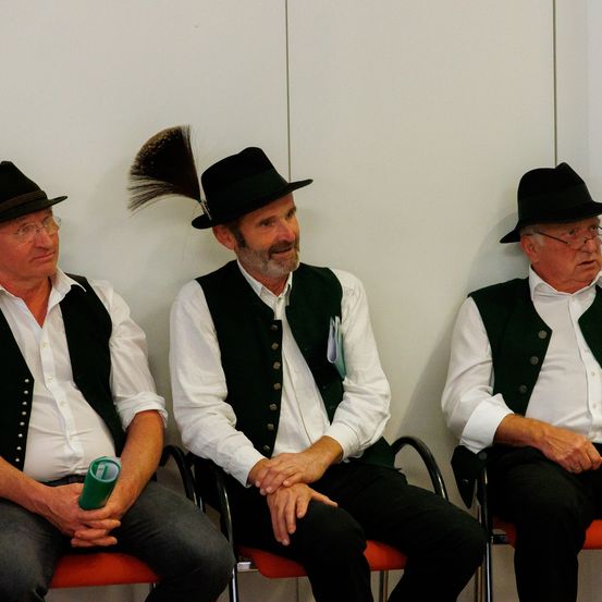 Drei ältere Männer in traditioneller Kleidung sitzen nebeneinander auf roten Stühlen an einer weißen Wand. Einer hat eine Feder in seinem Hut.