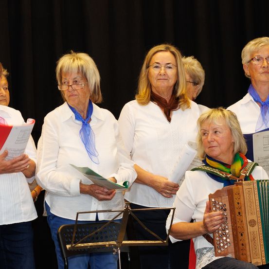 Eine Gruppe von Frauen, in Weiß gekleidet, steht auf einer Bühne. Eine Frau spielt Akkordeon, während andere Notenblätter halten. Sie scheinen sich auf eine Aufführung vorzubereiten.
