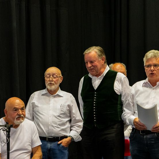 Fünf ältere Männer stehen auf der Bühne vor einem schwarzen Hintergrund. Einer hält ein Stück Papier, ein anderer ein Mikrofon. Alle tragen weiße Hemden, außer einem in einer grünen Weste.