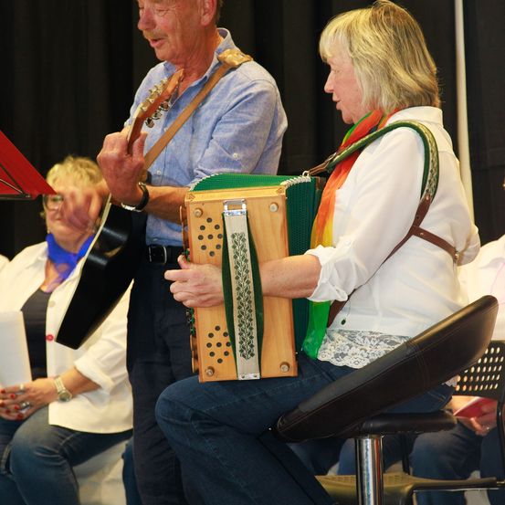 Ein Mann und eine Frau spielen Instrumente bei einer Aufführung, mit einer Frau im Hintergrund und einem schwarzen Vorhang als Hintergrund.