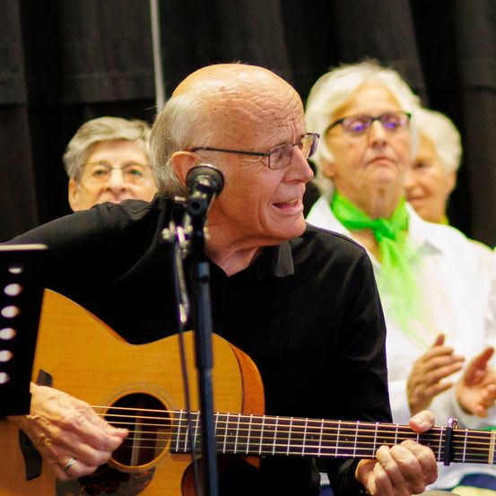 Ein älterer Mann spielt Gitarre und singt, während eine andere Frau klatscht und lächelt. Eine Frau mit grünem Schal schaut zu.