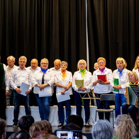 Ein Chor älterer Erwachsener steht auf der Bühne, hält Notenblätter und singt. Dahinter ist ein schwarzer Vorhang. Ein Publikum sieht zu.