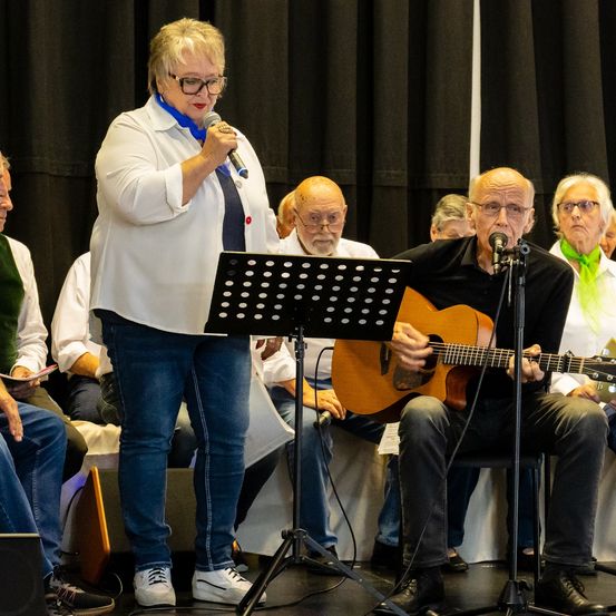 Eine Frau mit Brille und blauem Schal steht auf der Bühne und hält ein Mikrofon. Ein Mann mit Brille spielt neben ihr eine Gitarre. Dahinter sitzt eine Gruppe älterer Menschen auf Stühlen und schaut zu. Vor den Musikern steht ein Notenständer.