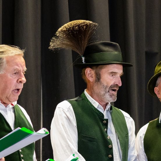 Drei ältere Männer stehen vor einem schwarzen Hintergrund. Der Mann in der Mitte singt und hält ein Buch. Der Mann auf der linken Seite singt und hält ein Buch. Der Mann auf der rechten Seite singt und hält ein Buch.