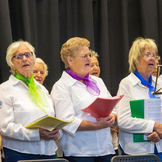 Eine Gruppe älterer Frauen in weißen Hemden und verschiedenen farbigen Schals singt auf der Bühne, hält Notenbücher und steht hinter einem Notenständer.
