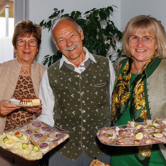 Drei Personen lächeln für ein Foto und halten Tabletts mit Essen. Der Mann in der Mitte trägt eine Weste mit Hirschmuster, während die Frauen Brillen und Schals tragen.