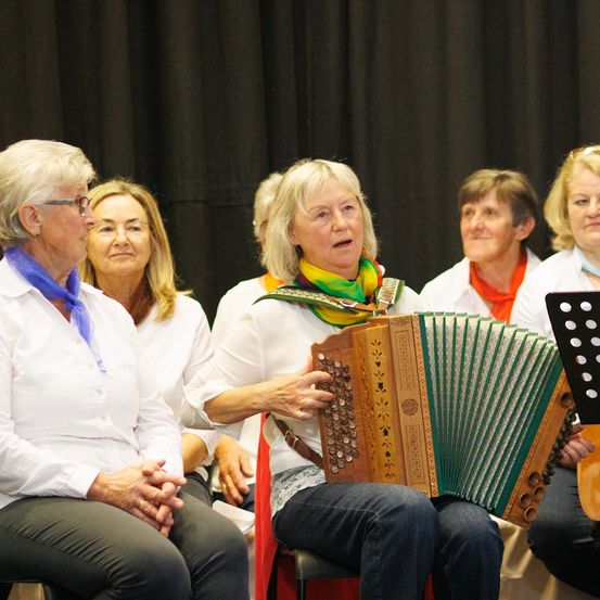 Eine Gruppe älterer Frauen sitzt auf der Bühne. Eine von ihnen spielt Akkordeon. Sie tragen alle weiße Hemden und blaue Schals. Hinter ihnen ist ein schwarzer Vorhang.