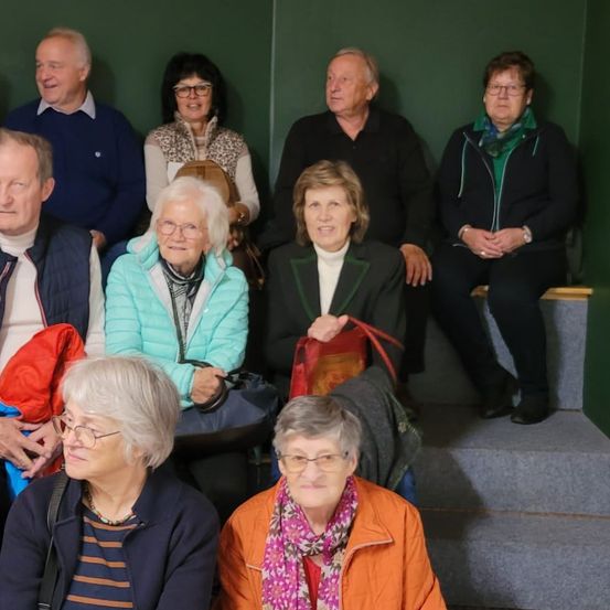 Eine Gruppe älterer Erwachsener, hauptsächlich Frauen, sitzen zusammen auf einer Bank, lächeln und schauen in die Kamera. Sie tragen Winterkleidung und einige haben Taschen auf dem Schoß. Sie befinden sich in einem Raum mit grünen Wänden.
