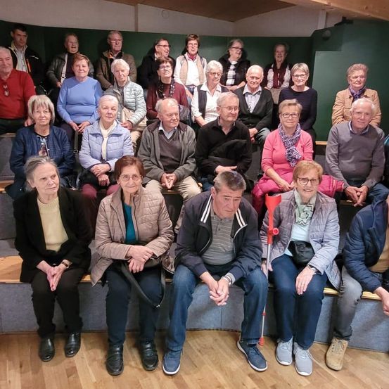 Eine Gruppe von Menschen sitzt zusammen auf einer Bank in einem Raum, einige mit Brillen und andere mit Accessoires wie Handtaschen und Gehstöcken.