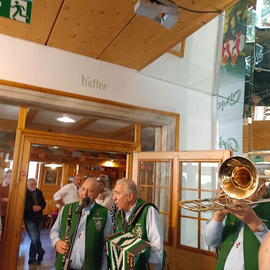 Zwei Männer spielen Musikinstrumente in einer Halle. Einer spielt Trompete, der andere Klarinette. Ein Schild mit der Aufschrift 'Kaffee' ist an der Wand zu sehen.