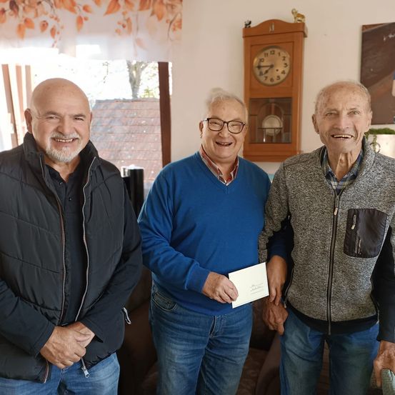 Drei ältere Männer stehen gemeinsam in einem Raum. Einer hält ein Papier. Sie alle lächeln. Hinter ihnen befindet sich eine Uhr, ein Gemälde und ein Fenster.