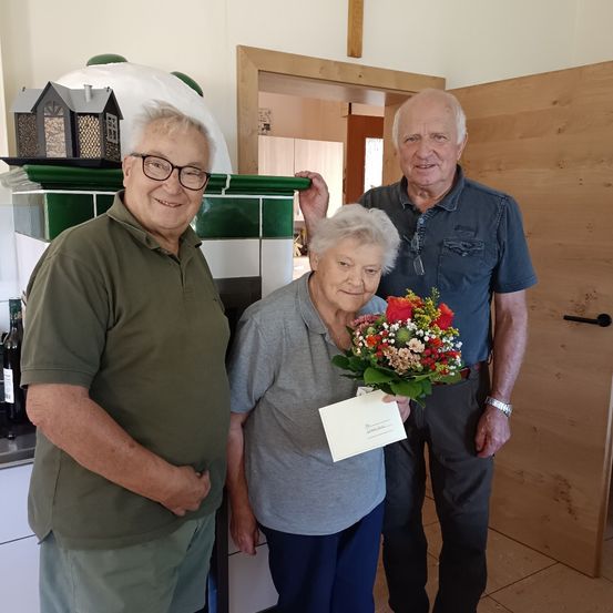 Drei Menschen stehen in einer Küche mit einer lächelnden älteren Frau, die Blumen und eine Karte hält, während ein Mann ihren Arm hält. Hinter ihnen befindet sich eine grün geflieste Wand und eine Holztür.